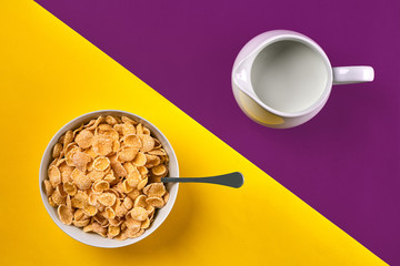 Bowl with corn flakes, jug of milk and spoon on purple and yellow background, top view