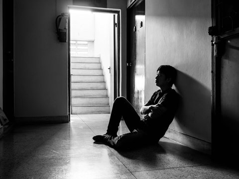 a sad young man sitting in the dark leaning against the wall with his back, The sun is in sorrow, black and white.