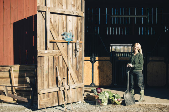 Female farmer with shovel standing by organic vegetables outside barn
