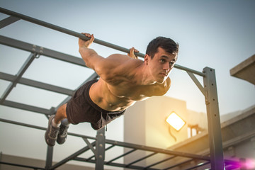 Young shirtless man doing exercises on horizontal bar outdoors. Calisthenics workout. - obrazy, fototapety, plakaty