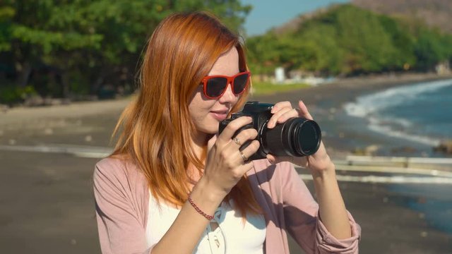 Young caucasian woman in sunglasses taking photo with reflex camera on volcanic sand beach in Pemuteran and smiling on sunny summer day