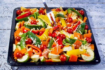 Mix of a raw colorful ecological summer vegetables, sprinkled with grated cheese, cooked for baked.  On a black metal baking sheet. Multi colored vegetarian food background. Top view.
