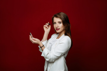 Beautiful woman model with long natural hair in a white t-shirt posing on red background.The girl makes up eyes with ink and looks in a small mirror. 