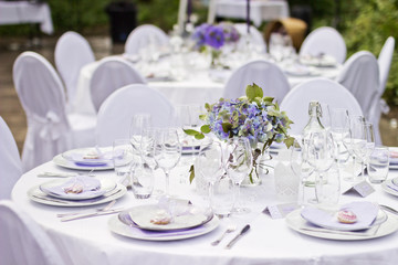 Outdoor wedding in Scandinavian style in old abandoned greenhouse. White tablecloth, white chairs, green trees in background. 