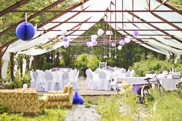 Outdoor wedding in Scandinavian style in old abandoned greenhouse. White tablecloth, white chairs, green trees in background. 