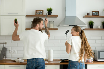 Funny couple having fun enjoying cooking in kitchen together, young man and woman preparing food feeling happy and playful helping standing their back to camera holding knife and spatula, rear view