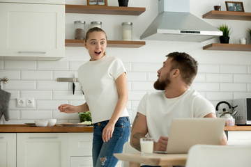 Obraz premium Surprised wife excited to hear good unbelievable news online from husband sitting with laptop at dining table, astonished happy woman distracted from cooking feeling amazed talking to man in kitchen
