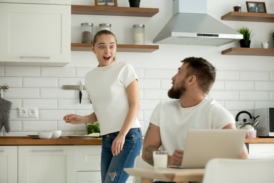 Surprised wife excited to hear good unbelievable news online from husband sitting with laptop at dining table, astonished happy woman distracted from cooking feeling amazed talking to man in kitchen