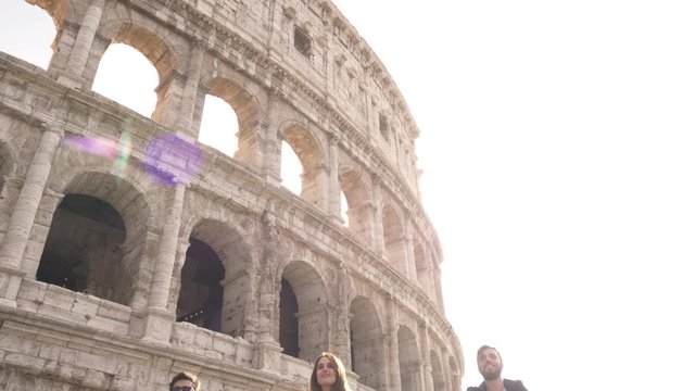 Three happy young friends tourists riding bikes with backpacks at Colosseum in Rome on sunny day slow motion camera steadycam ground shot