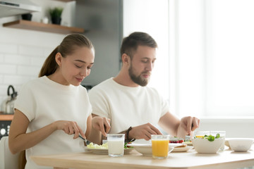 Obraz na płótnie Canvas Young happy couple eating with knife and fork together at home, man and woman having dinner at kitchen dining table, tasting healthy balanced delicious yummy breakfast, enjoying organic meal