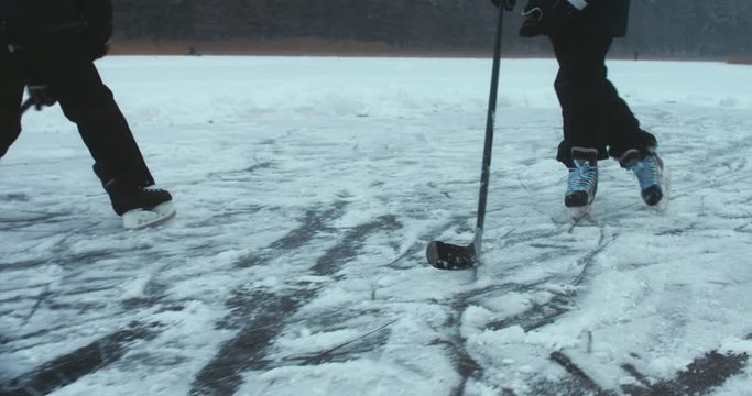 Father and son playing pond hockey on a frozen lake together, light snowfall. 4K UHD 60 FPS SLO MO