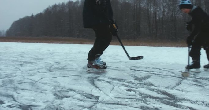 Father and son playing pond hockey on a frozen lake together, light snowfall. 4K UHD 60 FPS SLO MO