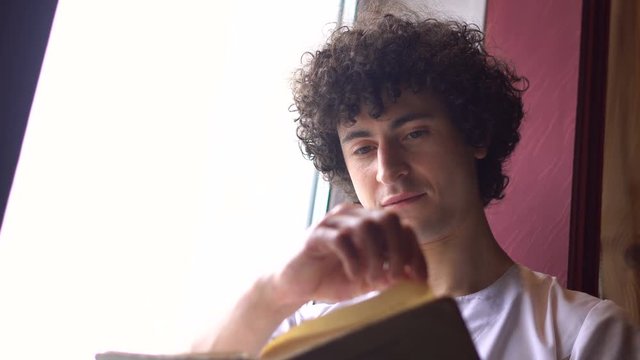 Young Curly Man in White Cloth is Sitting on Window Sill and Reading Book with Daylight from Window