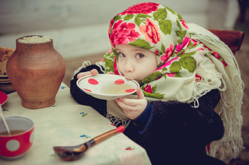 A little girl in an old fur coat and ornament scarf drinking tea at the holiday table. Shrovetide...