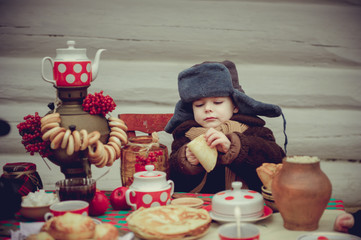 A little boy in an old fur coat and fur hat eats pancakes and jam at the holiday table. Shrovetide...