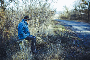 Naklejka na ściany i meble A young man sits on an old bench at a rural bus stop in the autumn time. The concept of waiting for a bus to a better life