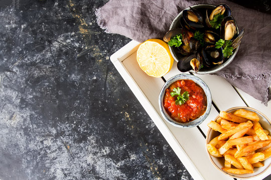 Mussels with herbs in a bowl with lemon and French fries on a white wooden board. Seafood. Food at the shore of the French Sea. Dark background. Top view