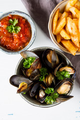 Mussels with herbs in a bowl with lemon and French fries on a white wooden board. Seafood. Food at the shore of the French Sea. Dark background. Top view