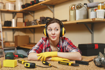 Pretty caucasian young brown-hair woman in plaid shirt, gray T-shirt, noise insulated headphones, yellow gloves working in carpentry workshop at wooden table place with piece of wood, different tools.