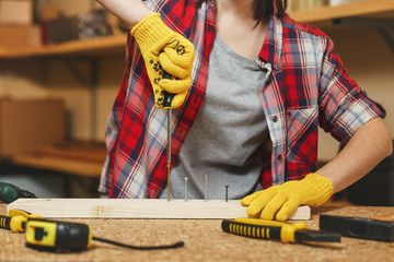 Close up of caucasian young woman in plaid shirt, gray T-shirt, yellow gloves twisting by screwdriver screw, working in carpentry workshop at wooden table place with piece of wood, different tools.
