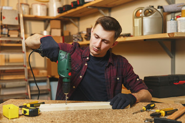 Handsome smiling caucasian young man in plaid shirt, black T-shirt, gloves drilling with power...