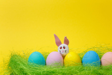 Row of five colorful pastel monophonic painted Easter eggs, white fun bunny in green dry grass on yellow background for card. Happy Easter concept. Copy space for advertisement. With place for text.