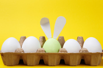 Row of white and light green painted Easter eggs with fun bunny ears in cardboard tray on yellow background for card. Happy Easter concept. Copy space for advertisement. With place for text.