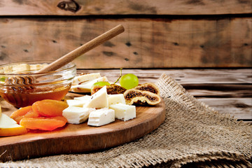 Cheese plate - various types of cheese, honey, grapes, dried apricots, nuts and figs on a wooden board on dark wooden background.