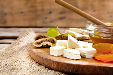 Cheese plate - various types of cheese, honey, grapes, dried apricots, nuts and figs on a wooden board on dark wooden background.