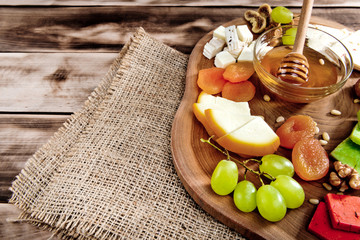 Cheese plate - various types of cheese, honey, grapes, dried apricots, nuts and figs on a wooden board on dark wooden background.