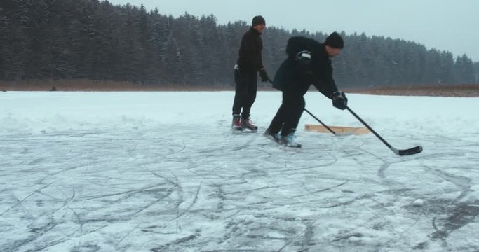 Two friends playing pond hockey on a frozen lake together, light snowfall. 4K UHD