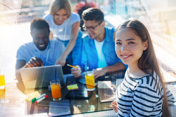 Perfect mood. Close up of pretty girl smiling at you while her friends using a laptop and having active discussion