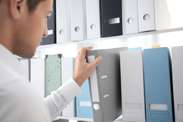 Young man taking folder with documents from shelf in archive