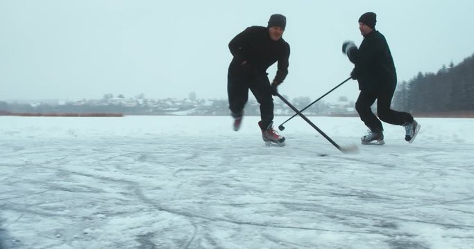 Two friends playing pond hockey on a frozen lake together, light snowfall. 4K UHD