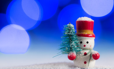 Cute Snowman christmas decoration, with beautiful blue and purple bokeh lights background, on top of white snow, holding a pine tree
