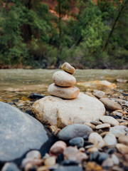 Fototapeta na wymiar Balanced pebble stone tower on river bed at river running through in Zion National Park in Utah, USA with river, trees and red rocks in background