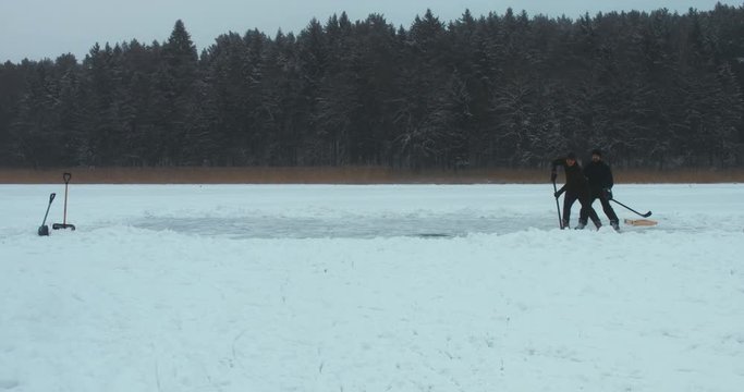 WIDE Two friends playing pond hockey on a frozen lake together, light snowfall. 4K UHD