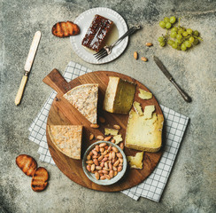 Flat-lay of cheese platter with cheese assortment, green grapes, bread, honey and nuts over grey concrete background, top view. Party or gathering eating concept