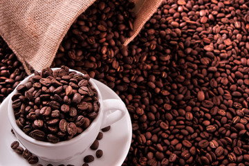 White coffee cup and saucer one single for espresso or capuccino filled full of coffee bean on a background of scattered dark coffee beans and bag sack  photo