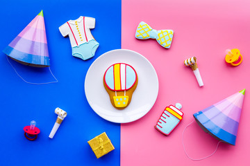 Celebrate child's birthday. Cookies in shape of baby accesssories, party hats, gift box, confetti on pink and blue background top view
