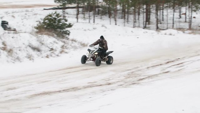 A young guy in a black hat and camouflage jacket extreme cornering snow track with the race on the ATV. Of the wheels flies the sand with the snow. Slowmotion.