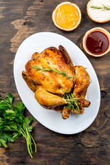 Roasted chicken with rosemary srved with sauces on white plate, wooden table, top view.