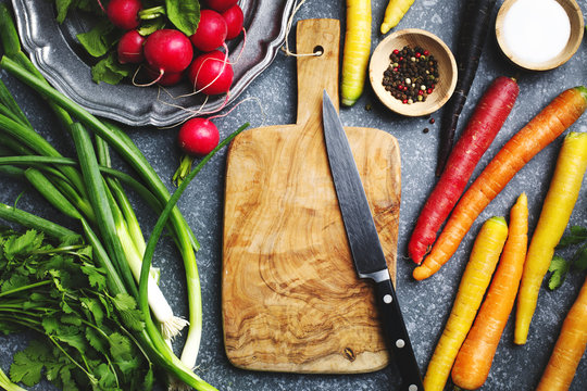 Fresh spring vegetables around wooden cutting board with kitchen knife for vegetarian cooking, top view