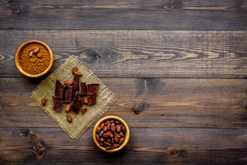 Make chocolate. Cocoa powder in bowl near cocoa beans and pieces of chocolate on dark wooden background top view copy space
