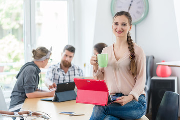 Portrait of a confident young woman holding a tablet and a coffee mug, while sitting on the desk of a co-working space for creative freelancers and entrepreneurs