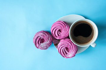 Pink marshmallows and a cup of coffee on a white saucer on a bright blue background. Place for text. Flat lay. 
