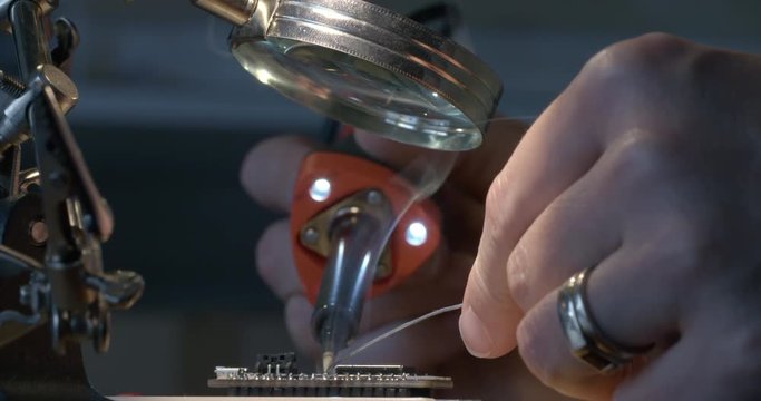 Hobbyists' hands soldering an electrical connection with a hot soldering iron fitted with LED lights. Smoking solder, looking across small circuit board. Recorded in 4K, slow motion at 60fps.