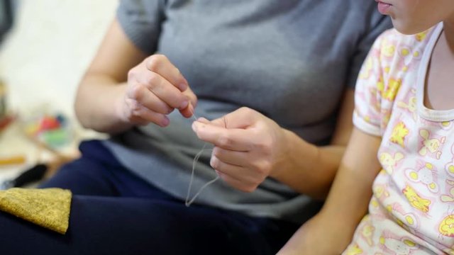 Mom teaches her daughter by sewing with a needle and thread. Mom and her little daughter sew clothes for a doll. The woman ties the end of the thread to the knot.