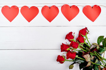 Bouquet of fresh red roses and garland of paper hearts on wood table, copy space. Greeting card mockup for Valentines day. Love concept. Top view, flat lay