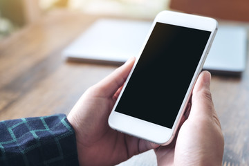 Mockup image of hands holding white smartphone with blank black desktop screen and laptop on wooden table